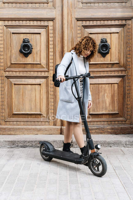 Full body of focus young African American female in coat with scooter standing on city street near historic building and looking down — Foto stock