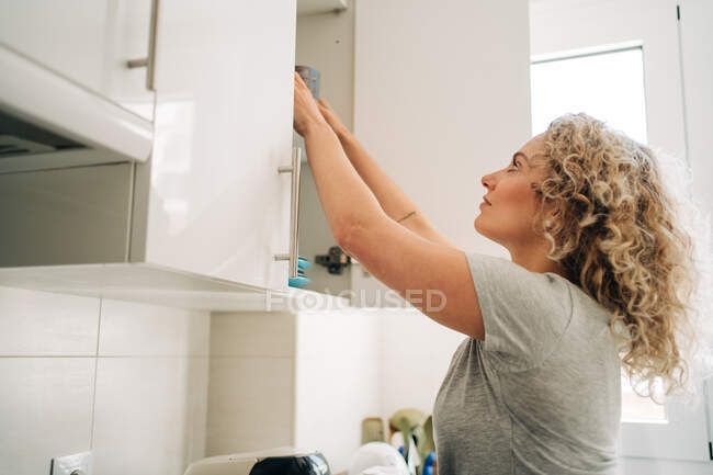 Junge positive Frau in lässigem Hemd, die Geschirr in den Schrank stellt, während sie in der hellen modernen Küche steht und nach oben schaut — Stockfoto
