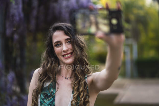 Joyeux magnifique femelle avec de longs cheveux bouclés prendre selfie sur téléphone portable tout en passant une journée chaude dans un parc abondant — Photo de stock