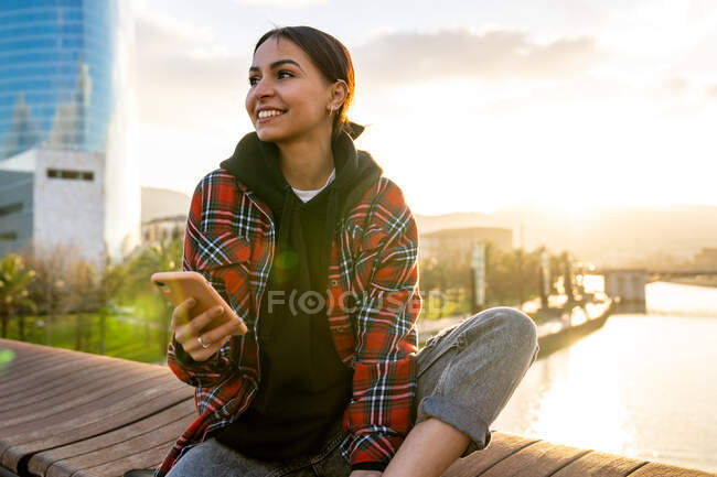 Jeune femme ethnique souriante en tenue décontractée messagerie texte sur téléphone portable sur pont au-dessus de la rivière sous un ciel nuageux à la lumière du soleil regardant loin — Photo de stock