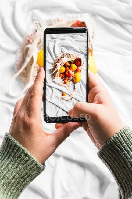 Overhead view of crop unrecognizable person touching screen on mobile phone while taking photo of fruits in zero waste bag — Stock Photo