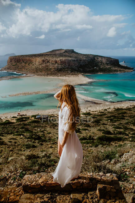 Бічний вид анонімної жінки в білому максі пляжі стоїть на кам'яному пагорбі і захоплюється мальовничим краєвидом пляжу Балос і довгим волоссям в сонячний спекотний день. — стокове фото