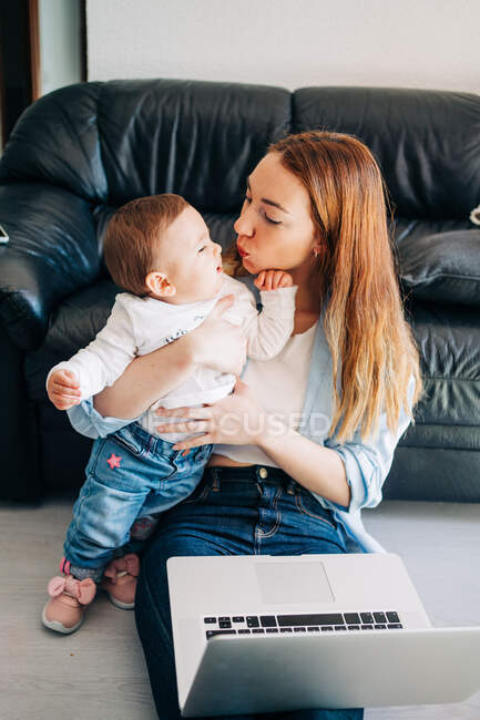 Jovem mãe vestindo roupas casuais abraçando bebê bonito e netbook de navegação enquanto sentados no chão juntos — Fotografia de Stock