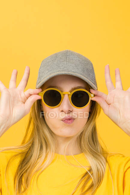 Young funny woman in yellow sweater and cap holding stylish sunglasses while standing looking at camera on yellow background — Stock Photo