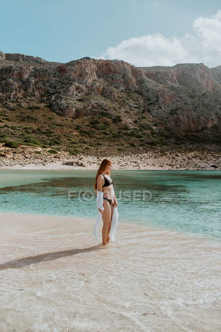 Ganzkörperschlanke Frau in schwarzem Badeanzug steht am Sandstrand von Balos vor felsigen Klippen und schaut bei klarem, sonnigem Wetter weg — Stockfoto