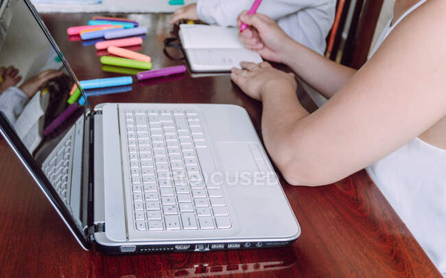 Cultivo mujer irreconocible y niño pequeño sentado a la mesa y dibujo con marcadores en el cuaderno - foto de stock