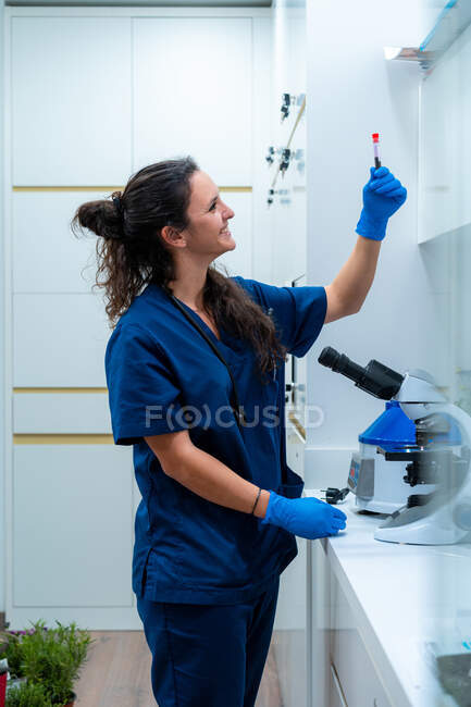 Visão lateral do conteúdo veterinário fêmea em uniforme com tubo de ensaio em pé à mesa com microscópio em laboratório — Fotografia de Stock