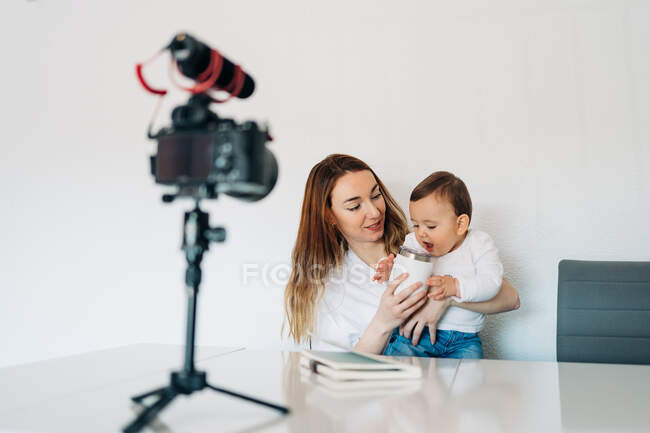 Позитивна молода жінка дає напій милій дитині, сидячи за столом і записуючи відео на камеру для особистого блогу — стокове фото