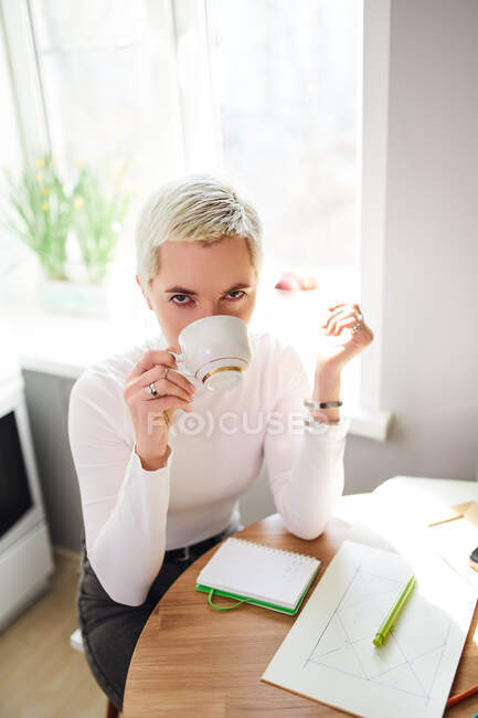 Female astrologist drinking hot beverage from cup while looking at camera at home in sunlight — Stock Photo