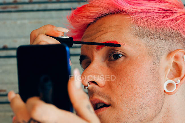 Homme homosexuel avec piercings et coupe de cheveux moderne appliquant mascara sur les cils avec applicateur contre téléphone portable — Photo de stock