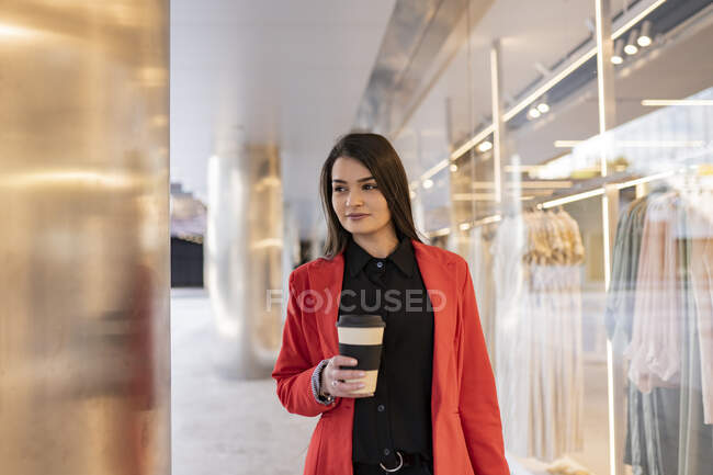 Весёлая женщина-покупатель с бумажными пакетами для покупок и кофе на вынос стоит рядом с витриной магазина в городе — стоковое фото