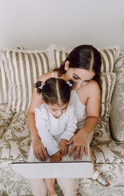 Dall'alto di donna etnica con bambina in ginocchio seduta in divano a casa e guardando cartoni animati su netbook insieme — Foto stock