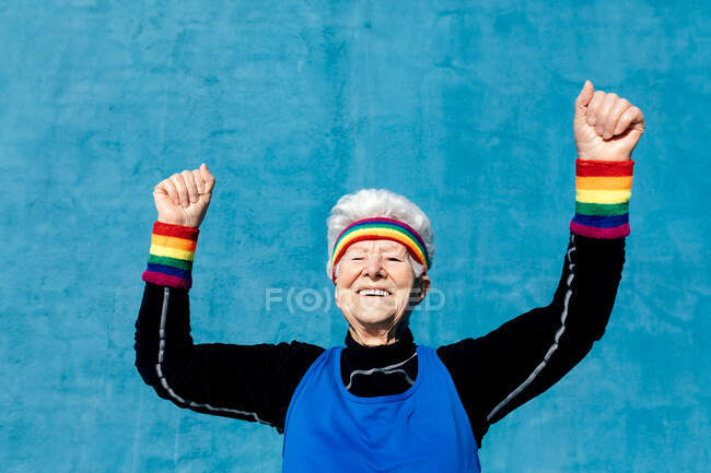 Emocionado sênior feminino em sportswear comemorando a vitória com punhos apertados e braços levantados em fundo azul no estúdio — Fotografia de Stock