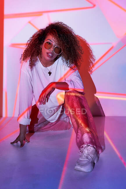 Full body attractive young African American female in stylish sunglasses sitting on floor and looking at camera in neon lights in dancing studio — Stock Photo