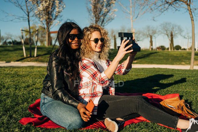 Вид збоку усміхнених багаторасових друзів, які сидять на ковдрі в парку і приймають власний портрет на старовинній миттєвій фотокамері — стокове фото