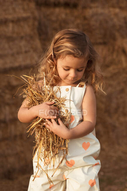 Side view of adorable child in overalls playing with hay near straw bales in countryside — Stock Photo