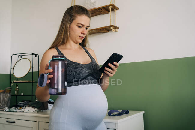 Молодая беременная женщина в спортивной форме смс-кой на мобильный телефон, стоя с бутылкой в комнате — стоковое фото