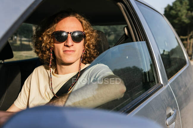 Serious young haired male in stylish sunglasses looking at camera through open window of car while sitting at driver seat — Foto stock