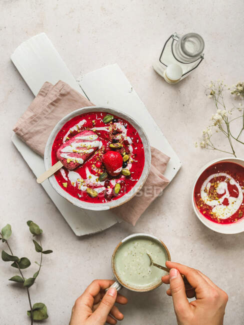 Top view of crop unrecognizable person stirring matcha tea in cup placed on table with sweet cold berry soup with popsicle in bowl — Stock Photo