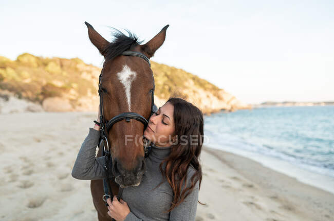 Young female kissing muzzle of chestnut stallion in bridle against wavy ocean — Stock Photo