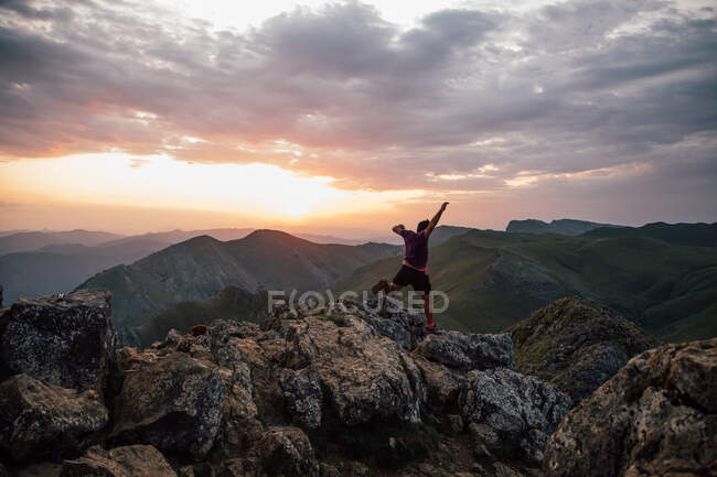 Вид ззаду людини, що стрибає через валуни високого піку гірського хребта під хмарним небом під час заходу сонця — стокове фото