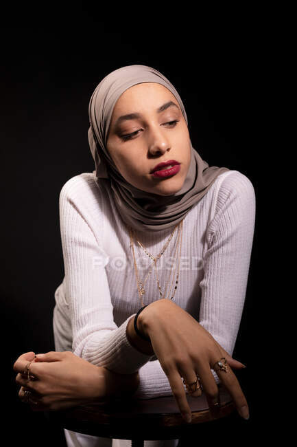 Modish mulher muçulmana confiante em hijab inclinado na cadeira e olhando para baixo no estúdio escuro — Fotografia de Stock