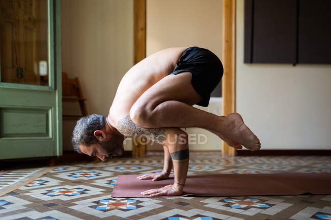 Вид сбоку сфокусированного гибкого мужчины, занимающегося йогой в Какасане, балансируя на коврике дома — стоковое фото