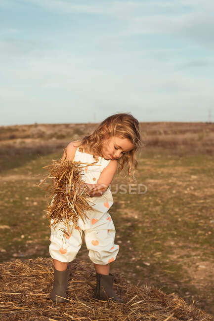 Adorable child in overalls playing with hay near straw bales in countryside — Stock Photo