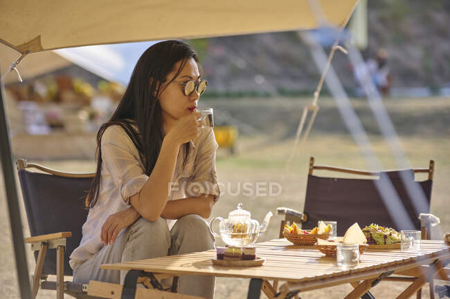 Hermosa mujer asiática étnica en gafas de sol sentada en la mesa bebiendo té mientras pasa un tiempo relajante en la zona de acampada durante las vacaciones - foto de stock