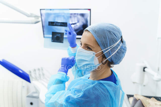 Жіночі ортодонти у стерильній формі та маску обличчя, що вказує на екран з рентгенівським зображенням зубів під час розмови з пацієнтом у клініці. — стокове фото