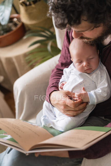 Bearded dad with adorable little child reading book while sitting with crossed legs in armchair in house — Stock Photo