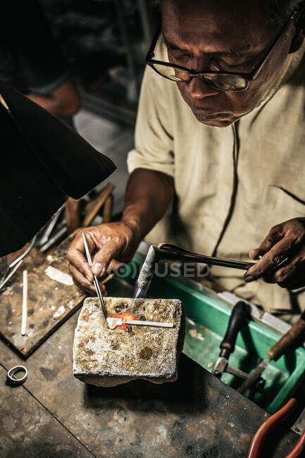 Fro acima maduro etnia homem com queima de tocha aquecimento pedaço de metal trabalhando na mesa em oficina de artesanato — Fotografia de Stock