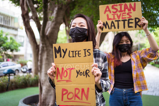 Mujeres étnicas con máscaras sosteniendo carteles protestando contra el racismo en la calle de la ciudad y mirando hacia otro lado - foto de stock