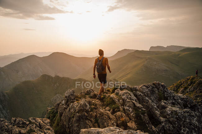 Vista posterior del hombre anónimo con mochila caminando sobre la cima rocosa de la montaña en el valle contra el cielo del atardecer - foto de stock