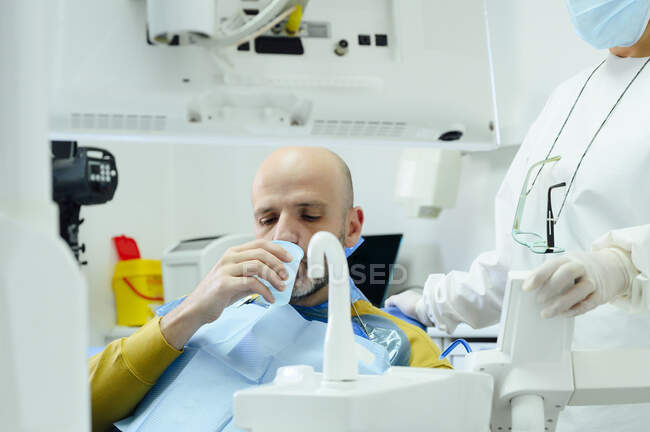 Врожай невизначеної жінки-стоматолога в уніформі і стерильній масці проти людини, що заливає воду з крана в скло після пероральної операції — стокове фото