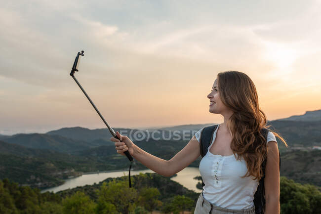 Vista lateral de la mujer viajera con la mochila de pie en la colina y tomando uno mismo tiro en el teléfono inteligente en el fondo de la cordillera en verano - foto de stock