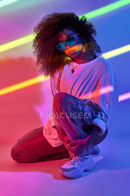 Full body stylish confident African American female dancer with curly hair and sunglasses standing in neon lights in dancing studio — Stock Photo