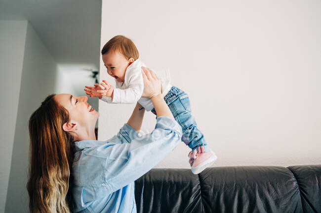 Side view happy young mom in casual shirt raising cute laughing baby on hands while spending time together in light living room — Stock Photo