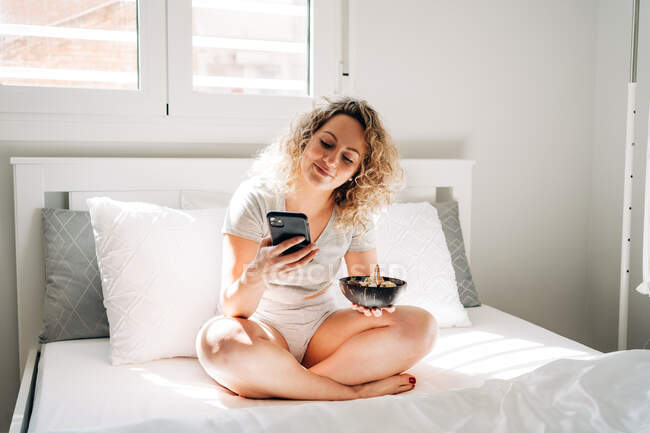 Ganzkörperfreudige junge Frau in häuslicher Kleidung isst leckeres Frühstück in der Schüssel, während sie morgens mit überkreuzten Beinen auf dem gemütlichen Bett sitzt und auf dem Handy surft — Stockfoto