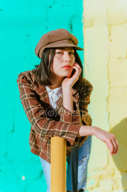 Young ethnic female in trendy apparel touching face while leaning forward and looking at camera in sunlight - foto de stock