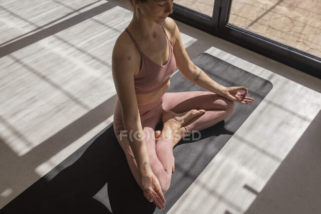 Desde arriba de la cosecha femenina concentrada en ropa deportiva sentada con las piernas cruzadas y los ojos cerrados en la esterilla de yoga en casa - foto de stock
