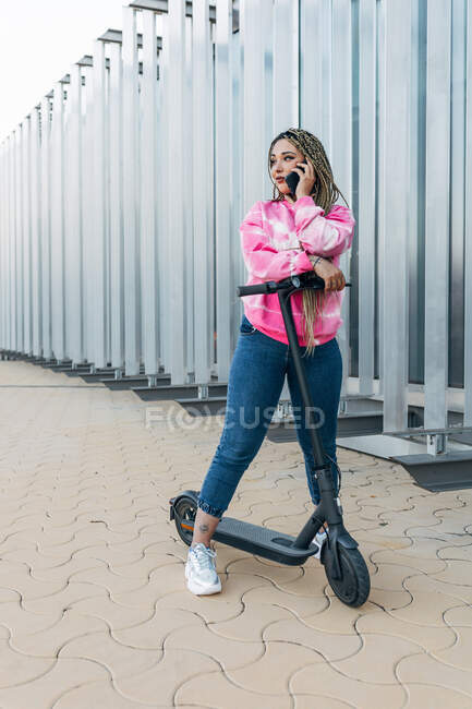 Молода жінка в повсякденному одязі з електричним скутером розмовляє на мобільному телефоні, дивлячись на міську прогулянку — стокове фото