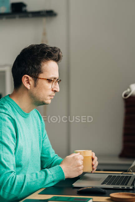 Side view of male distance worker in eyeglasses with mug of hot drink against netbook in house — Stock Photo
