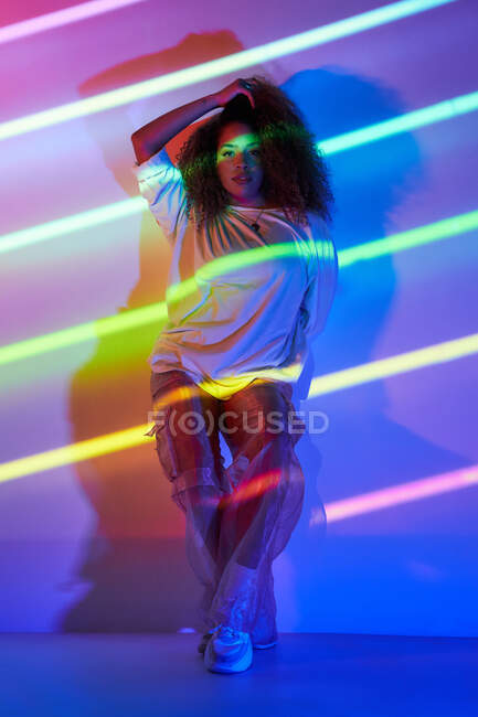 Полностью уверенная в себе афро-американская танцовщица с вьющимися волосами и солнцезащитными очками, стоящая в стене и смотрящая на камеру в неоновом свете в танцевальной студии — стоковое фото