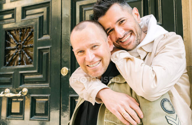 Cheerful young homosexual diverse men in stylish outfits smiling and embracing while standing on street near door and looking at camera — Stock Photo