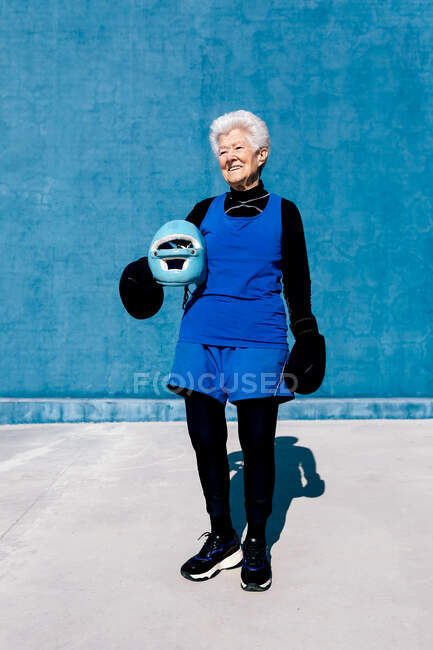 Mujer madura sonriente de cuerpo entero en ropa deportiva y guantes de boxeo de pie con casco en la mano contra la pared azul y mirando hacia otro lado - foto de stock