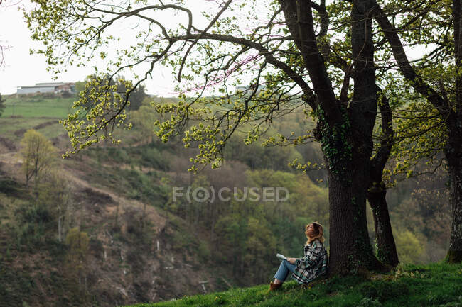 Side view of female traveler with paper guide sitting on meadow between green trees and mount in countryside — Foto stock