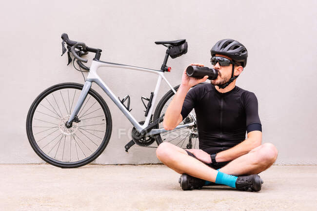 Male cyclist in sportswear with bottle sitting with crossed legs against bike on walkway — Foto stock