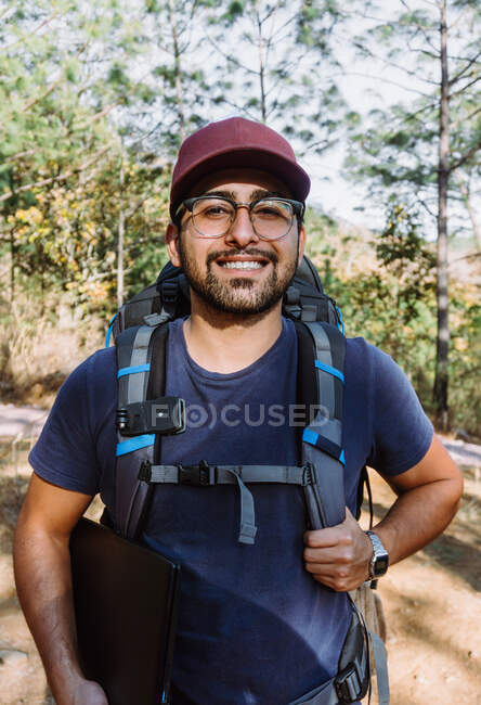 Bearded male backpacker in cap walking among trees and plants in woods in sunny day — Stock Photo