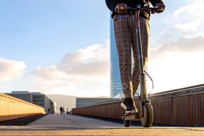 Засеянный неузнаваемый этнический предприниматель, катающийся на электрическом скутере по дорожке на городском мосту против зданий под облачно-голубым небом — стоковое фото